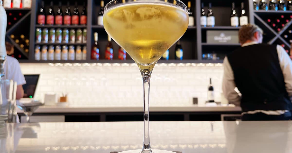 A coupe of champagne on a white bar counter, shelves of bottles behind