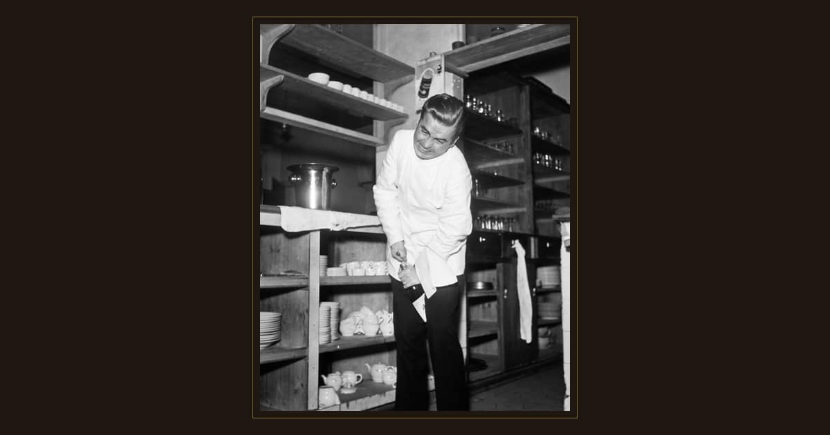Black-and-white photo of a waiter in a white jacket opening a wine bottle in a restaurant pantry, 1954