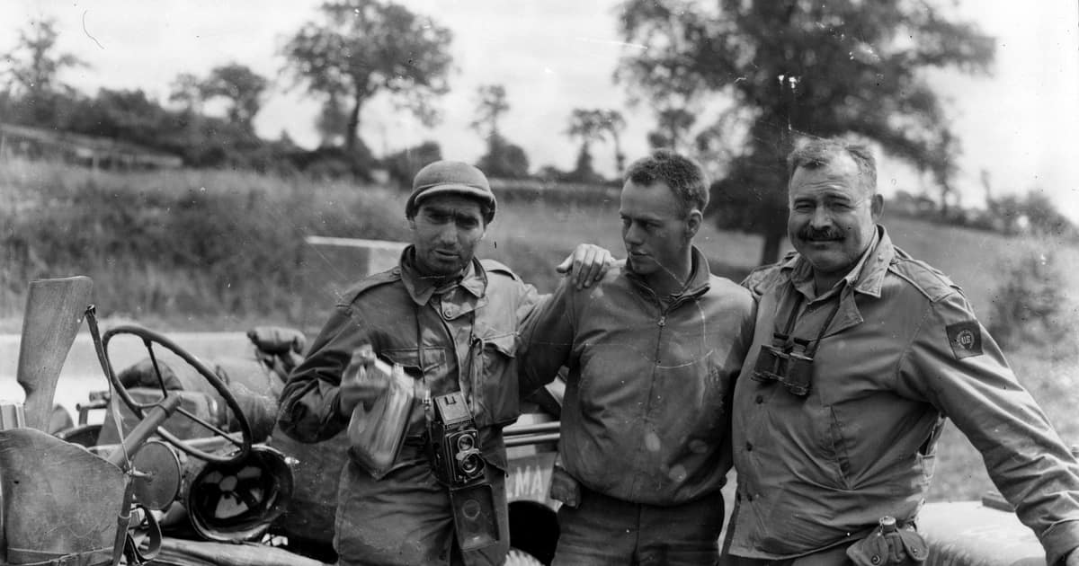 Black-and-white photo of Ernest Hemingway and Robert Capa standing by a jeep with an American soldier, 1944